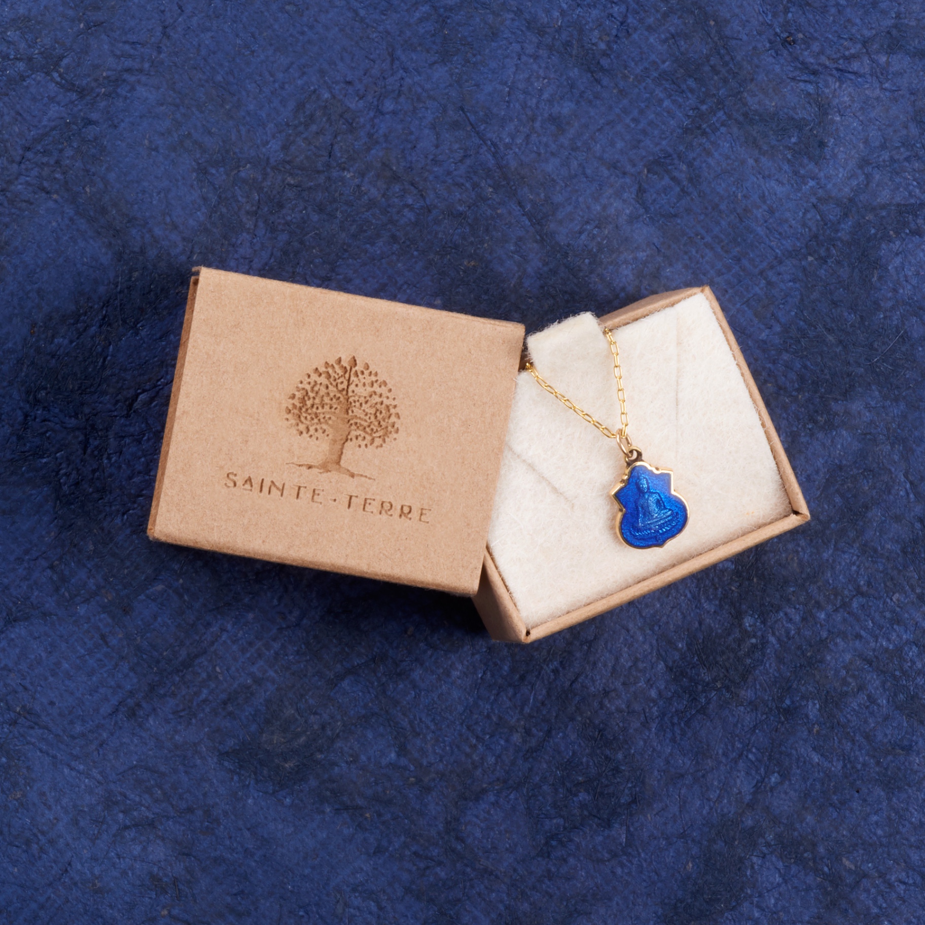 The small recycled kraft box, engraved with the Sainte-Terre tree logo, holding the Petite Buddha medallion