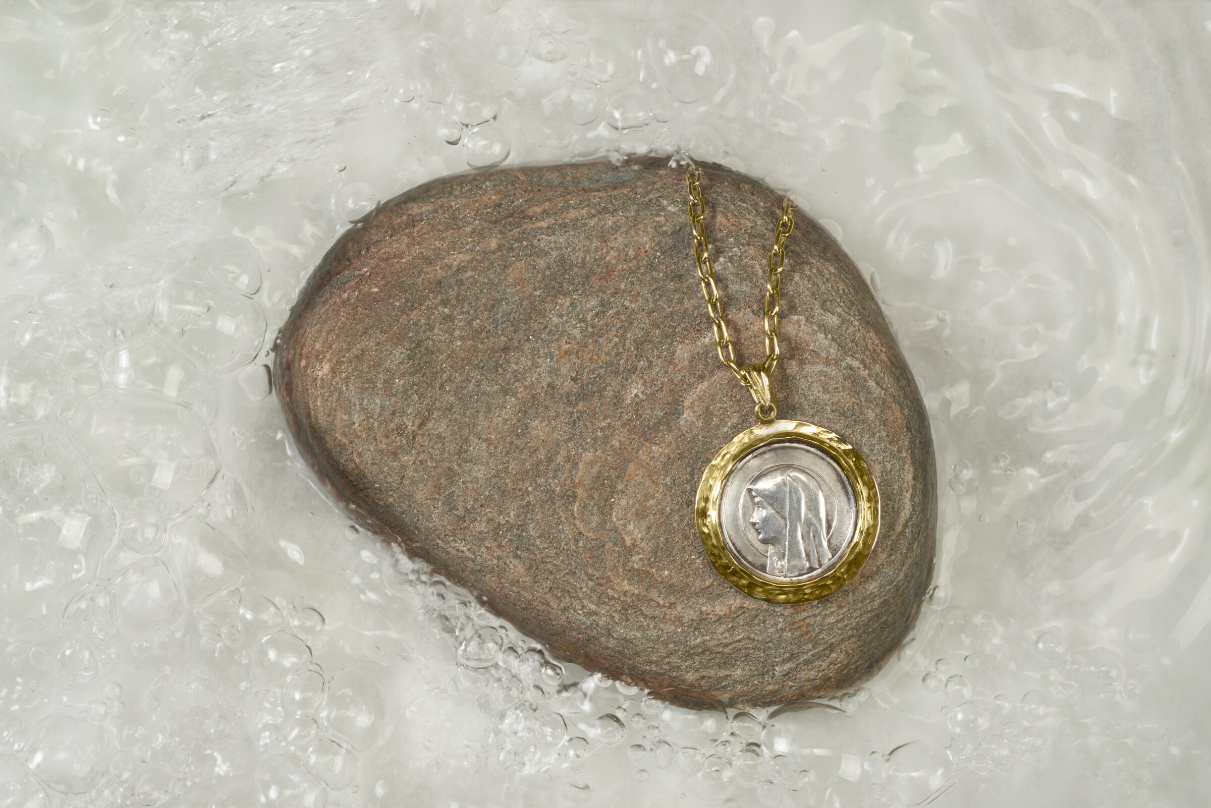 Our Lady of Lourdes medallion in silver and 14k gold, resting on a river stone in flowing water